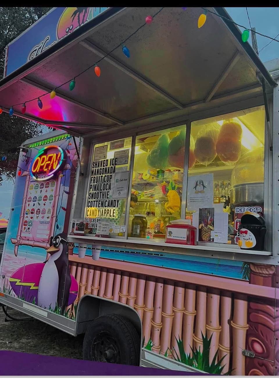 A shaved ice truck lit up at night with its serving window open and string lights overhead, at Coyoteville in Alice, Texas